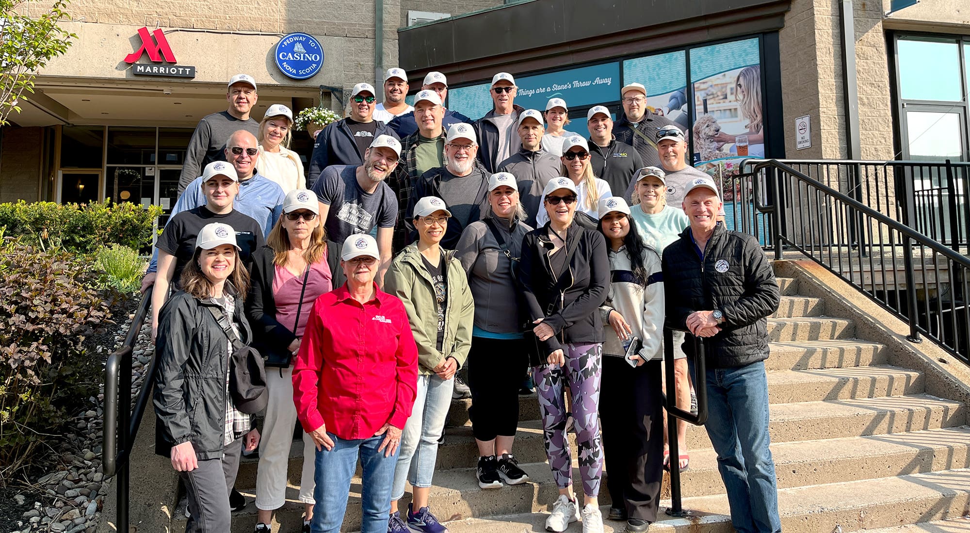 A group of people in Halifax gather on outdoor steps in front of a Marriott hotel, wearing matching white caps as they prepare for a charity walk supporting Homes for Heroes.